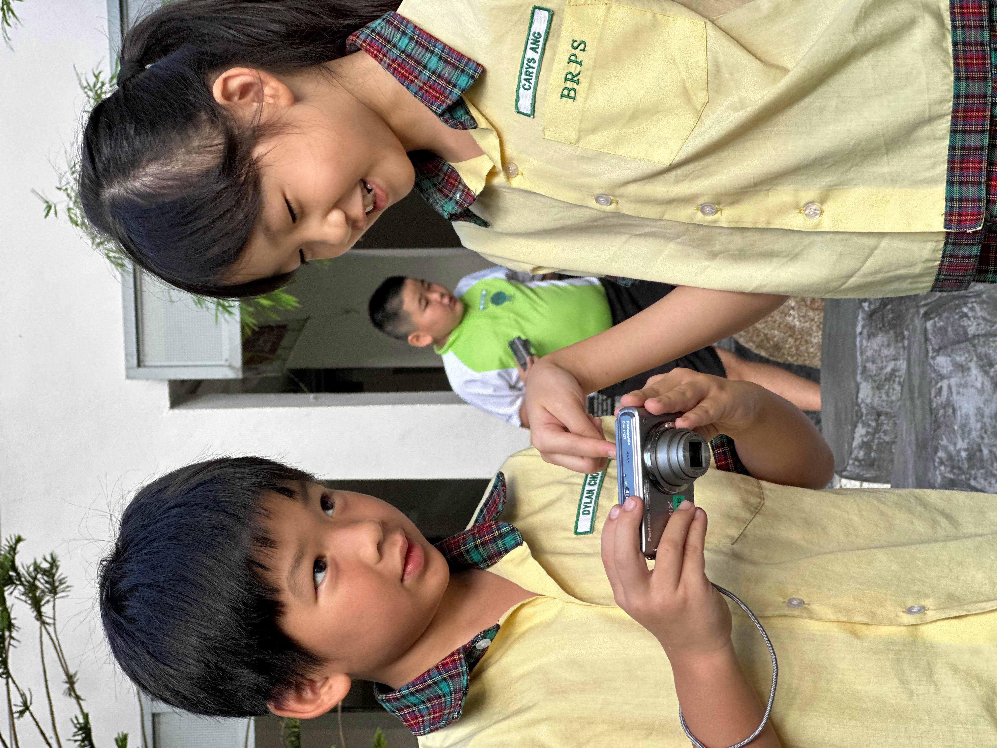 Two children in yellow school uniforms look at a digital camera while a man stands behind them.
